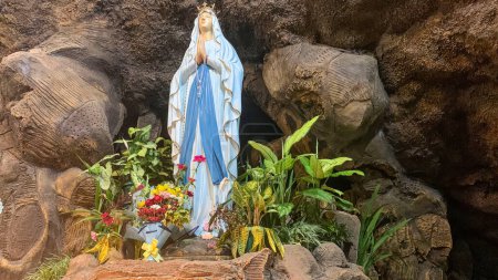 Foto de Estatua de la Santísima Virgen María en la Iglesia Católica Romana, en la cueva de la Virgen María, en una capilla cueva de roca Iglesia Católica con flores tropicales alrededor en el asiático - Imagen libre de derechos