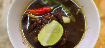 Foto de Rawon o sopa de carne de vacuno negro indonesio, de color negro son de la nuez indonesia llamado kluwek. Se sirve con lima, pasta de chile, huevo salado y galletas tempe. Perfecto para recetas, artículos o cualquier contenido de cocina o blog - Imagen libre de derechos