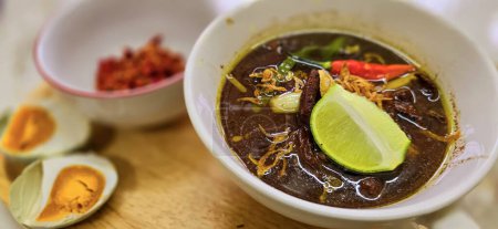 Rawon or Indonesian black beef soup, black color are from indonesian nut called kluwek. Served with lime, chili paste, salted egg, and tempe cracker. Perfect for recipe, article, or any cooking contents or blog
