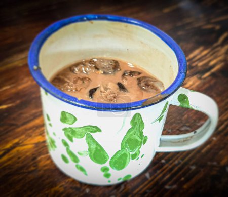 Ice chocolate drink on a vintage Indonesian iron mug with random green pattern called Blirik cup or cangkir Blirik on bokeh background along with chocolate filling bread on a cafe in Bogor