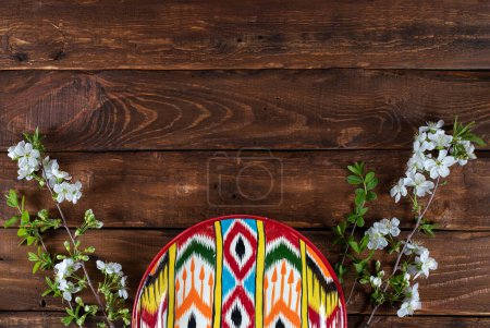Rishtan Uzbek national plate and  blooming cherry flowers on wooden background. Space for text. Vertical adras pattern