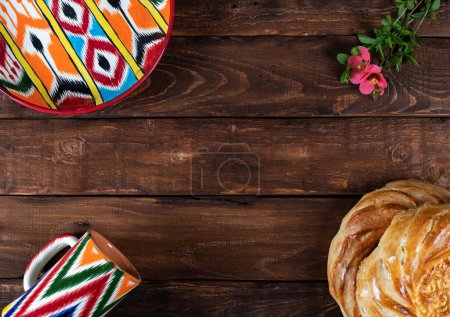 Photo for Rishtan Uzbek national plate and mug, journey cake bread and pink flowers on wooden background. Postcard for Navruz, Kurban bayram - Royalty Free Image