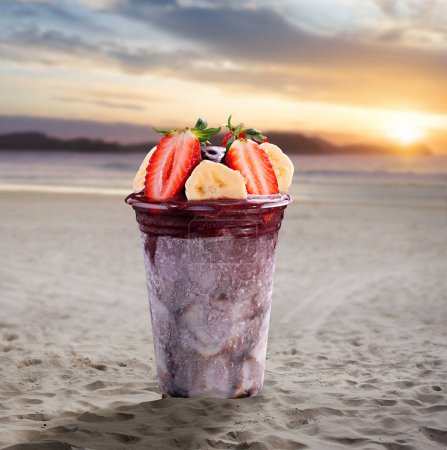 Foto de A cup of acai topped with strawberry and banana on a beach at sunset - Imagen libre de derechos