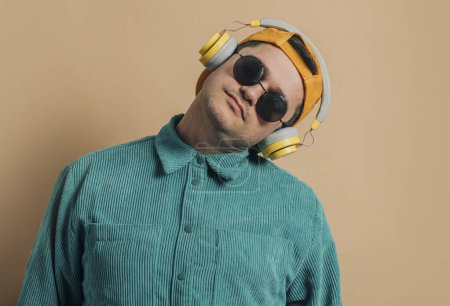 Téléchargez les photos : Stylish caucasian man in green shirt, headphones, sunglasses and cap on brown background - en image libre de droit