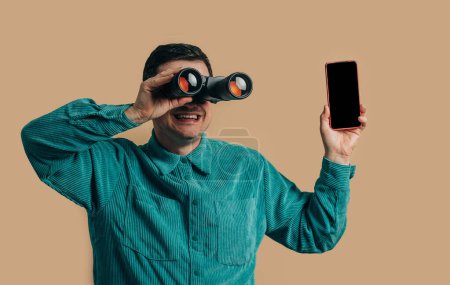 Téléchargez les photos : Stylish caucasian man in green shirt with smartphone and binoculars on brown background - en image libre de droit