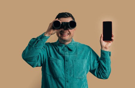 Téléchargez les photos : Stylish caucasian man in green shirt with smartphone and binoculars on brown background - en image libre de droit