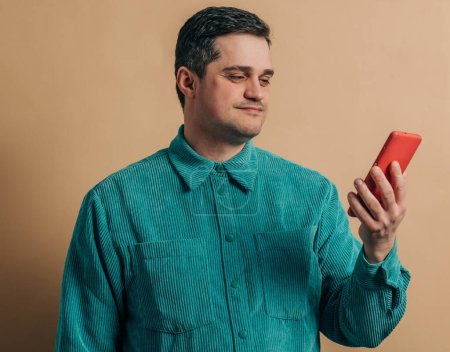 Foto de Elegante hombre caucásico en camisa verde con teléfono inteligente sobre fondo marrón - Imagen libre de derechos