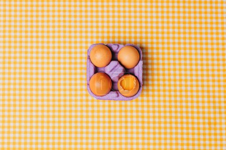 Photo for Four chicken eggs in purple egg box on yellow plaid tablecloth, top view - Royalty Free Image
