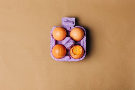 Téléchargez les photos : Quatre œufs de poulet dans une boîte à œufs violette sur fond éco-cuir marron, vue du dessus - en image libre de droit