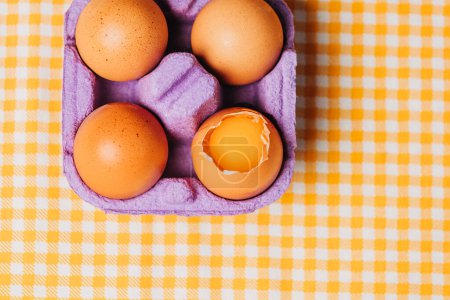 Foto de Cuatro huevos de pollo en caja de huevo púrpura en mantel a cuadros amarillo, vista superior - Imagen libre de derechos