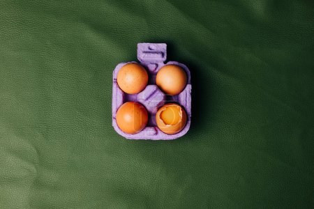 Téléchargez les photos : Quatre œufs de poulet dans une boîte à œufs violette sur fond éco-cuir vert, vue du dessus - en image libre de droit
