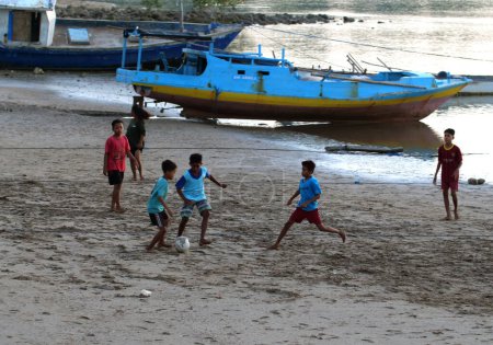 Téléchargez les photos : Maumere, Flores, Indonésie- 23 mai 2023 : certains enfants jouent au ballon sur la plage avec des bateaux en arrière-plan - en image libre de droit