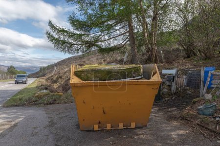 A bright yellow coloured skip with a mattresson the top which is covered in moss