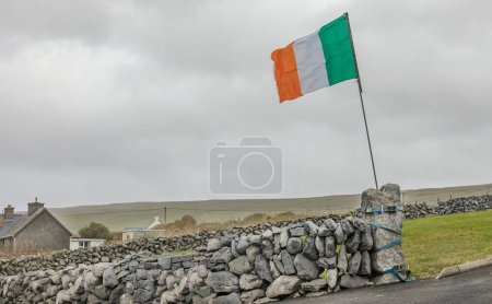 Irish flag flapping in the breeze attached to a gate post at the end of a stone wall