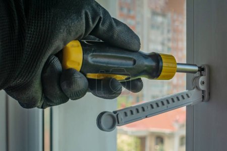 Photo for Male using screwdriver tightens screw that fixes plastic window opening limiter Close-up of gloved hand with screwdriver tightening window stopper fixing screw onto plastic window frame. Home repairs. - Royalty Free Image
