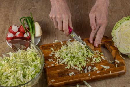 Photo for A woman uses a kitchen knife to cut fresh white cabbage on a wooden cutting board. Close-up female hands chopping cabbage for making summer salad. The concept of cooking food in the home kitchen. - Royalty Free Image