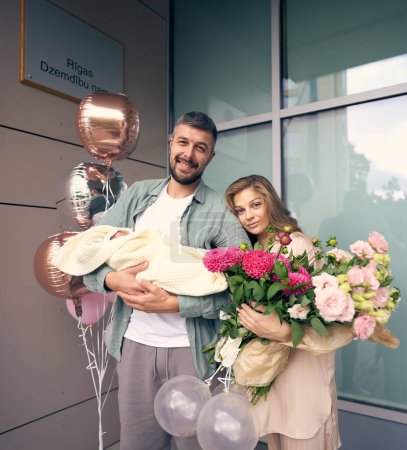 Téléchargez les photos : Happy caucasian couple with newborn baby. Father holds baby and mother holds a large bouquet of different bright flowers. Translation: Maternity home in Riga - en image libre de droit