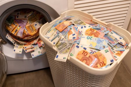 A big pile of money in an open washing machine. The big basket full of money is in front of. Money laundering concept.