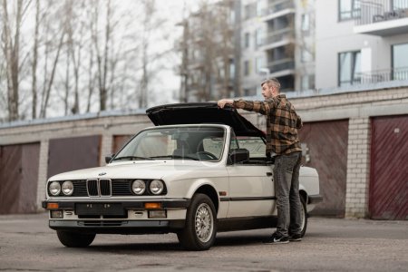 Photo for Riga, Latvia - April 1, 2024: A man folding the roof of white classic convertible BMW E30 car. Old garages and modern building in the background. - Royalty Free Image