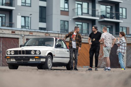 Photo for Riga, Latvia - April 1, 2024: A car owner shows to the group of young people his white classic convertible BMW E30 car. Old garages and a modern building in the background. - Royalty Free Image