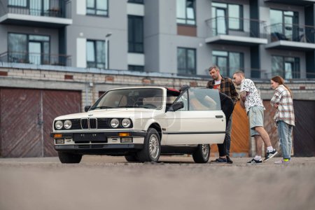Photo for Riga, Latvia - April 1, 2024: A group of young people looking with interest into white classic convertible BMW E30 car. Old garages and a modern building in the background. - Royalty Free Image