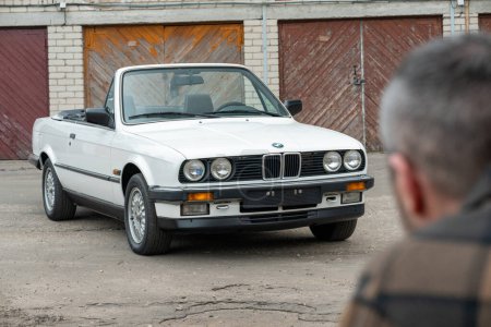 Photo for Riga, Latvia - April 1, 2024: A man looking on his white classic convertible BMW E30 car with opened roof. Old garages in the background. - Royalty Free Image