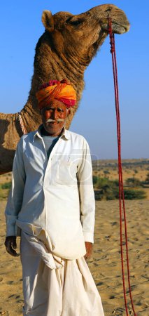 Foto de DESERT THAR RAJASTHAN INDIA - 02 13 2023: Camaleero indio (conductor de camellos) con camellos en dunas del desierto de Thar. Jaisalmer, Rajastán, India - Imagen libre de derechos