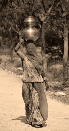 Foto de RURAL RAJASTHAN INDIA - 02 13 2023: Vida tribal pueblo vida mujeres llevando agua en macetas en cabeza escasez de agua de verano - Imagen libre de derechos