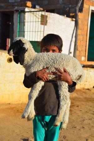 Téléchargez les photos : THAR DESERT JAISALMER RAJASTHAN INDIA - 02 13 2023 : Petit garçon portant son petit mouton. Les enfants ont la responsabilité de prendre soin de leur maison et de leur famille ; ils vont chercher de l'eau et broient du grain pour se nourrir. - en image libre de droit