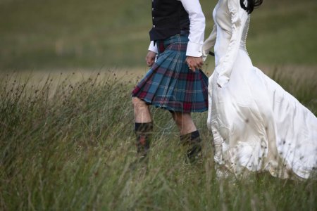 Téléchargez les photos : Un couple écossais nouvellement marié se promène dans un champ herbeux à Glencoe, dans les hautes terres écossaises. Il portait un kilt écossais et elle portait une robe de mariée blanche - en image libre de droit