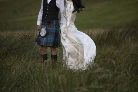 Photo for A newly married Scottish couple strolls through a grassy field in Glencoe, the Scottish highlands. He dressed in a Scottish Kilt and she dressed in a white wedding dress - Royalty Free Image