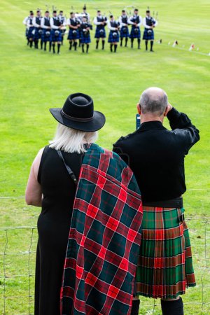 Foto de Un hombre y una mujer vestidos de negro. Él lleva un kilt y ella lleva un chal escocés. La pareja fotografía un desfile de bandas de pipa escocesas durante los Juegos Escoceses de las Tierras Altas en Crieff - Imagen libre de derechos