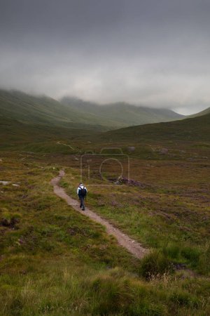Ein Mann wandert an einem Tag mit dramatischem Himmel vom Glencoe Lookout auf einem Bergpfad in den Glencoe Mountains
