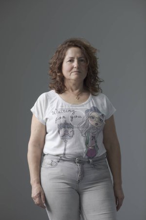 Portrait with white background of an adult woman with curly brown hair. White short-sleeved t-shirt with drawings and gray jeans. I would be