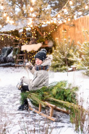 Foto de Lindo chico con ropa de invierno sosteniendo una linterna mientras está sentado en un trineo con un pino afuera. Humor de Navidad - Imagen libre de derechos