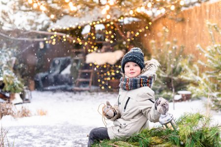 Foto de Niño con ropa de invierno sosteniendo una linterna mientras está sentado en un trineo con un pino afuera. Humor de Navidad - Imagen libre de derechos