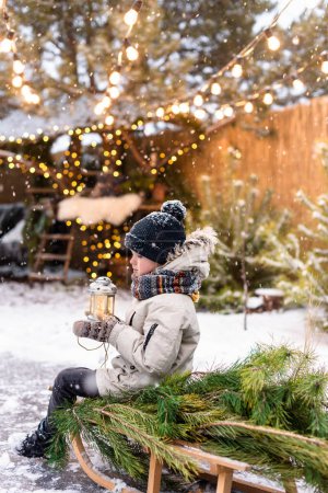 Foto de Niño con ropa de invierno sosteniendo una linterna mientras está sentado en un trineo con un pino afuera. Humor de Navidad - Imagen libre de derechos