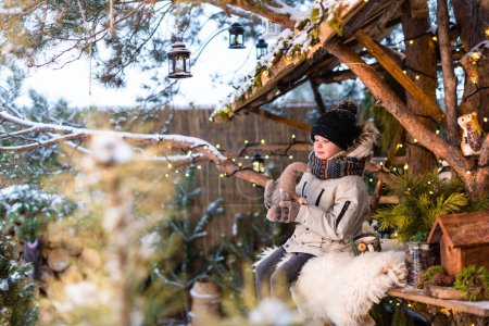 Foto de Un niño lindo en ropa de invierno se sienta en un pino en una casa entre la decoración de Navidad - Imagen libre de derechos