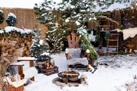 Foto de Decoración de Navidad en el patio: un árbol de Navidad con una guirnalda, sillas de madera junto al fuego, leña, coronas de Navidad, casa del árbol para el niño, una guirnalda colgante de linternas, - Imagen libre de derechos