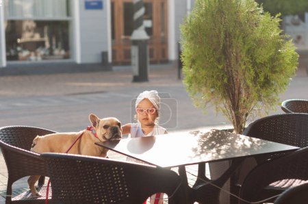 Little cute girl with french bulldog in a chair on the terrace of a cafe in the middle of the city