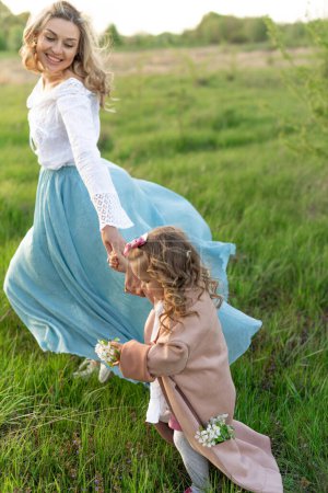 Una joven madre de pelo rubio con una larga falda azul camina con su pequeña hija en medio de un prado verde