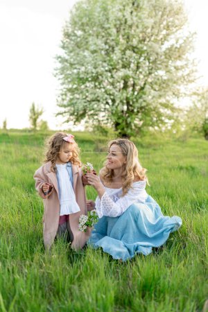 Foto de Una joven y hermosa madre y su pequeña hija están recogiendo flores en el prado - Imagen libre de derechos