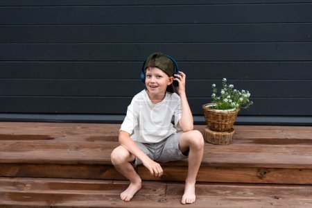 Funny and smiling boy in headphones on a wooden terrace near a flowerpot with copy space