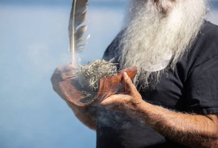 Téléchargez les photos : Les mains humaines tiennent un plat en bois avec des branches de plantes australiennes et une plume sur fond bleu, le rite rituel de la fumée lors d'un événement communautaire autochtone en Australie - en image libre de droit
