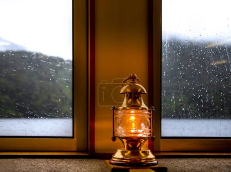 Eine Laterne mit warmgelbem Licht vor dem Hintergrund eines verregneten Fensters in einer Schiffskabine, ein Symbol für Komfort und Sicherheit, gemütliches Bild