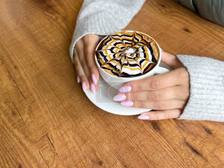 Photo for Girls hands hold white cup of coffee with spider web pattern on top of foam, against background of wooden table, close-up, top view - Royalty Free Image