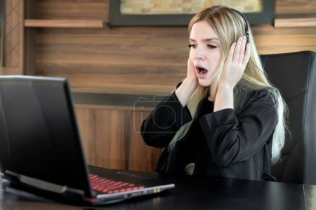 Photo for Young business woman sitting in front of laptop with expression of shock on her face with her mouth open and holding face with hands - Royalty Free Image