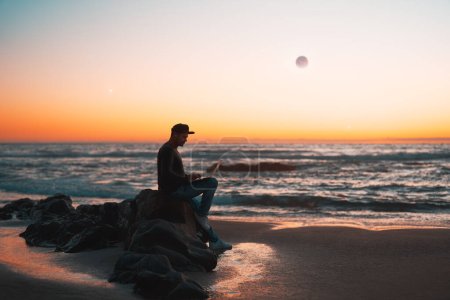Silhouette einer Person, die zur goldenen Stunde an ihrem Laptop im Freien am Strand arbeitet, Profilansicht