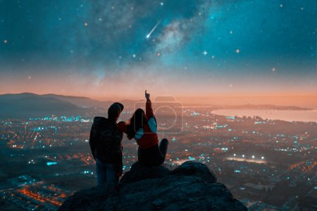 Foto de Siluetas de una pareja sentada en la cima de la montaña mirando y señalando la estrella fugaz y la Vía Láctea sobre las luces de la ciudad en el horizonte - Imagen libre de derechos