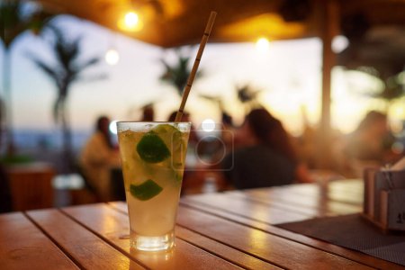 glass cup with cold caipirinha and straw on a wooden table in a bar at sunset on the beach of Ipanema	